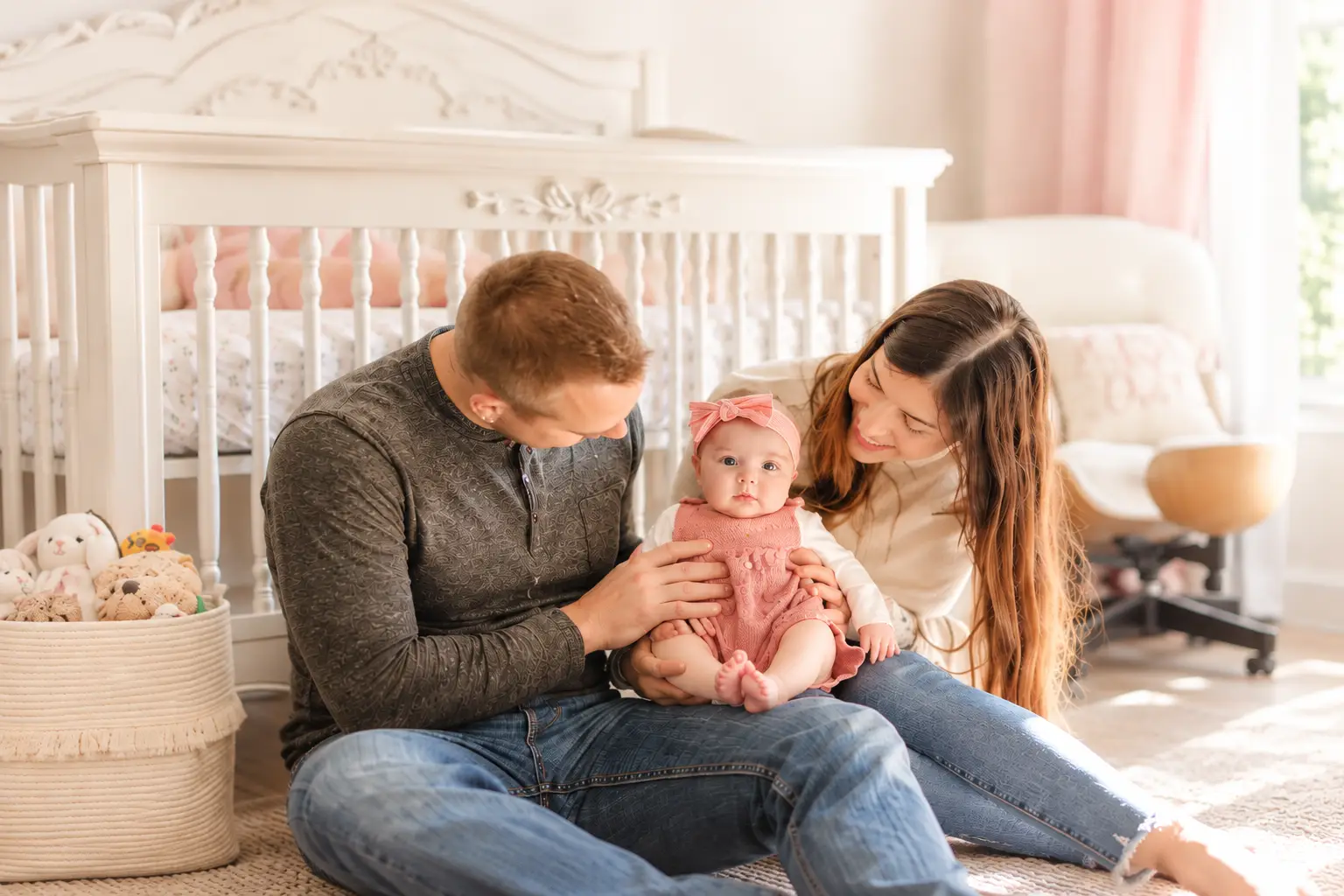 Smiling parents sit on the floor with their baby girl in a pink outfit in a bright nursery.