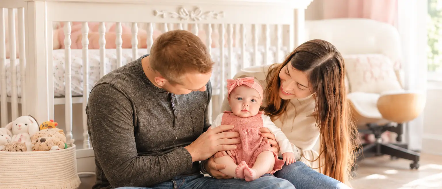 Smiling parents sit on the floor with their baby girl in a pink outfit in a bright nursery.