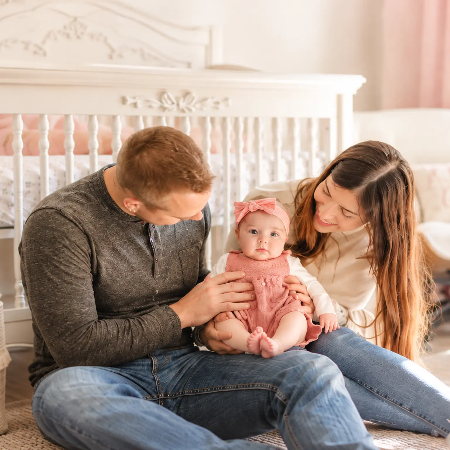 Smiling parents sit on the floor with their baby girl in a pink outfit in a bright nursery.
