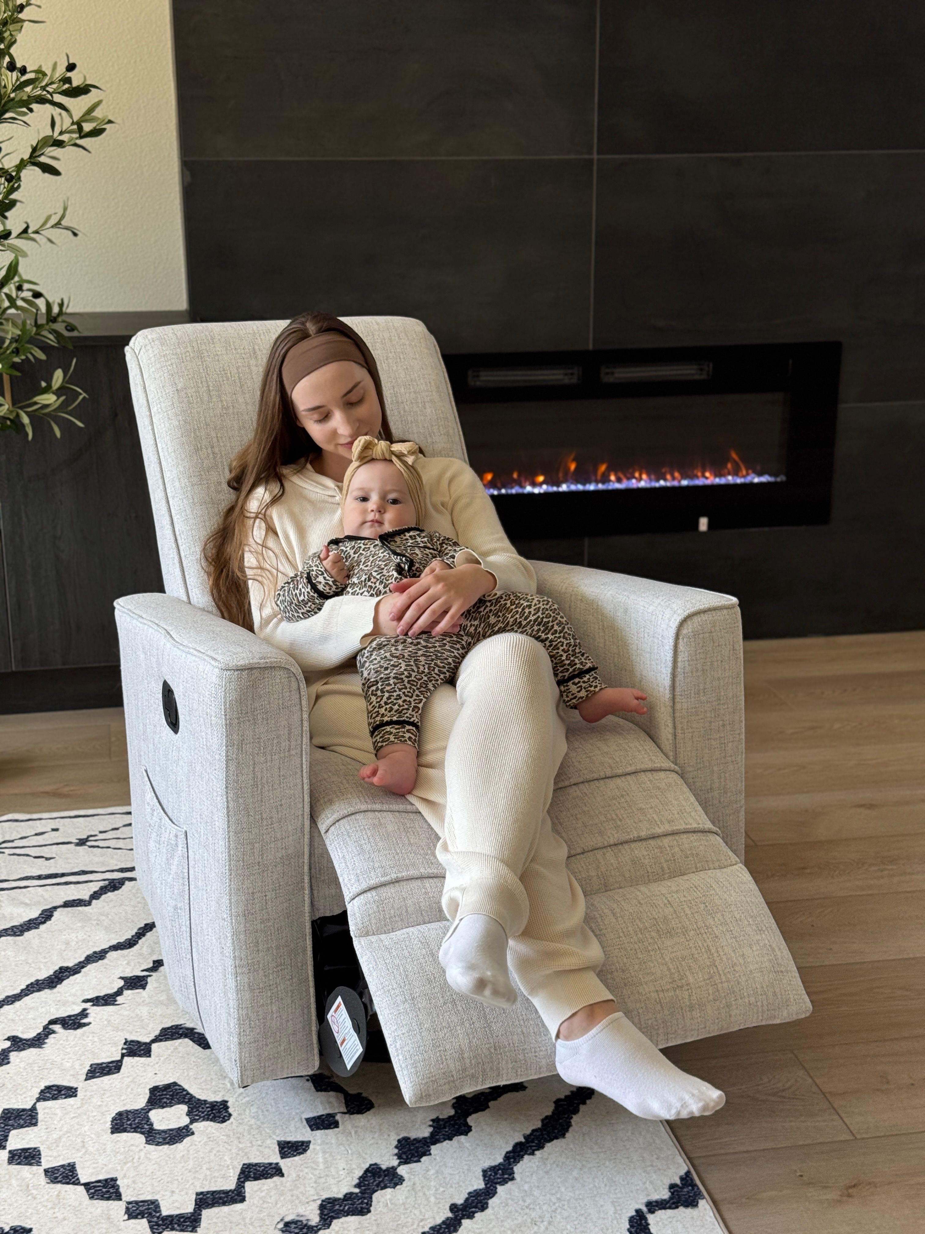 Mother sitting on Evolur Raleigh recliner chair in fog color holding baby in cozy living room with fireplace