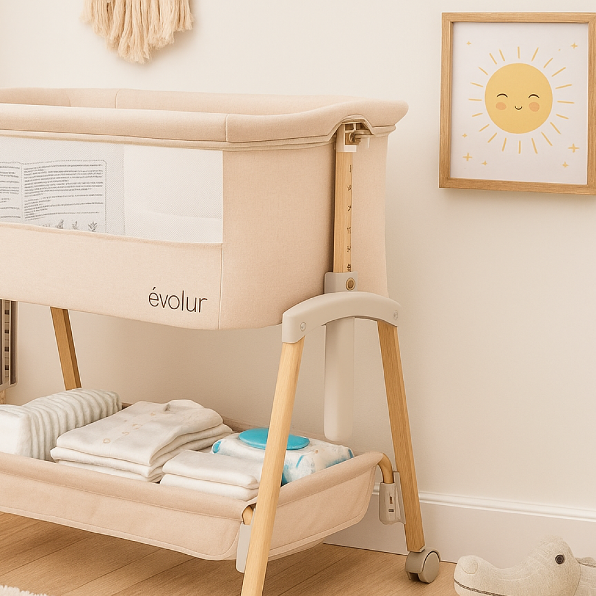 Evolur Lullaby bassinet with wooden legs and mesh sides, styled in a cozy nursery with folded baby clothes underneath, a smiling sun wall art, and a plush crocodile toy on the floor.