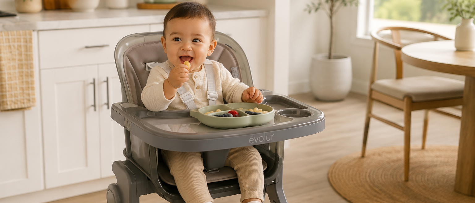 Happy baby in a gray high chair eating fruit from a green divided plate.