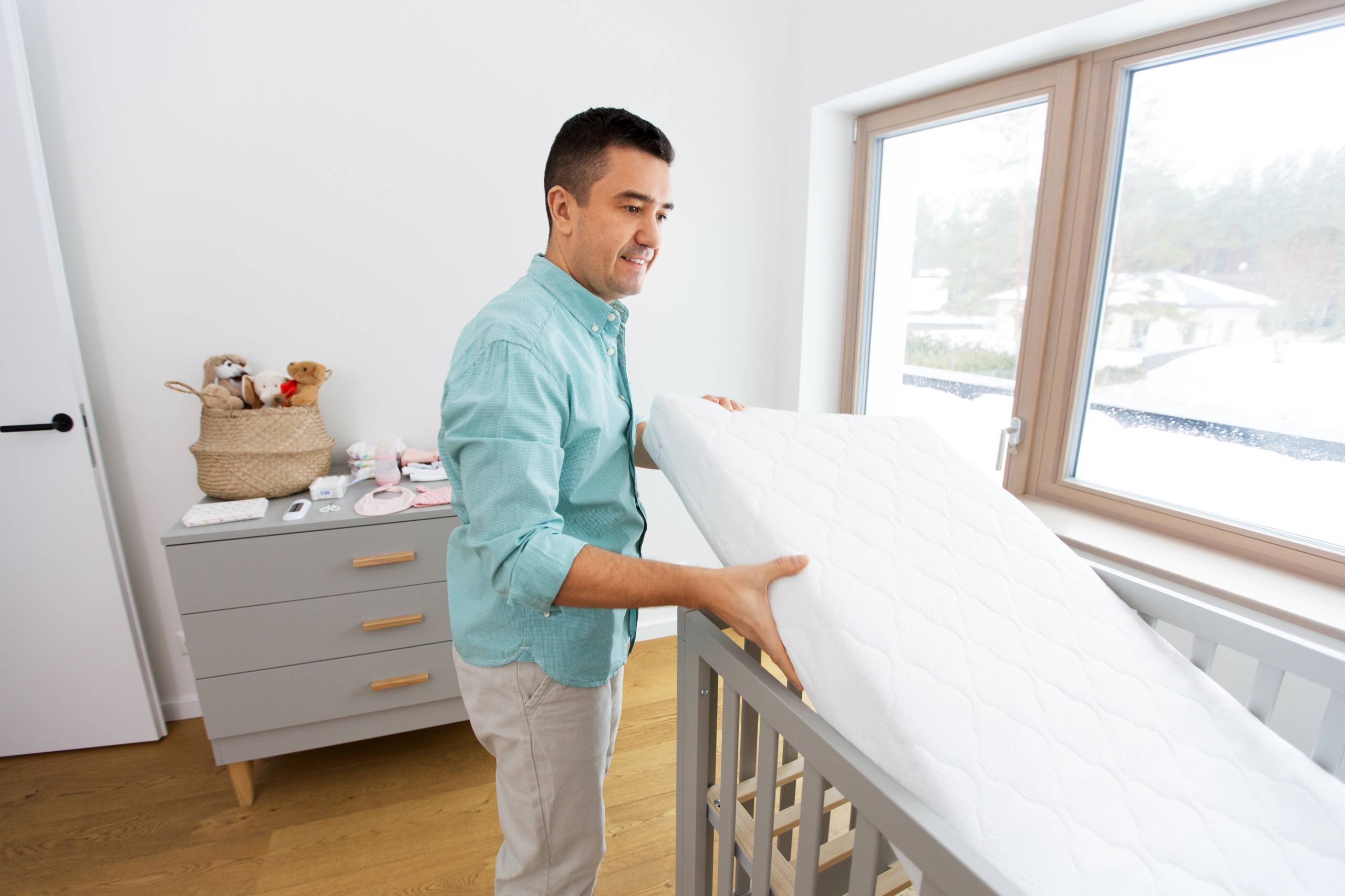 Father placing a white Mattress into a gray crib in a bright, modern nursery