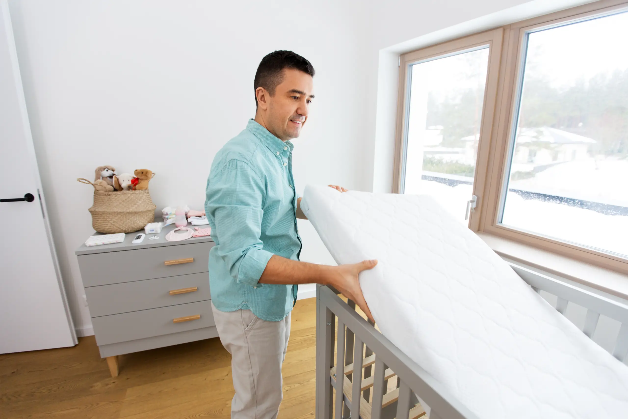 Father placing a white Mattress into a gray crib in a bright, modern nursery