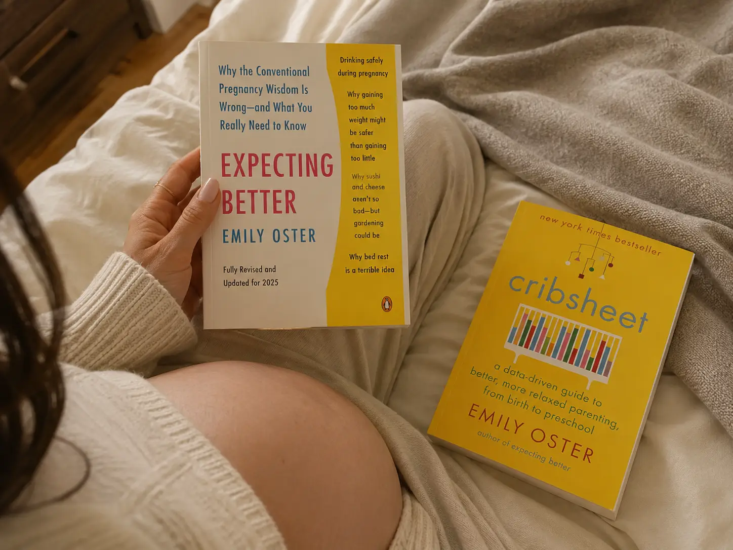 A pregnant person reads "Expecting Better" by Emily Oster, with "Cribsheet" by the same author lying nearby.