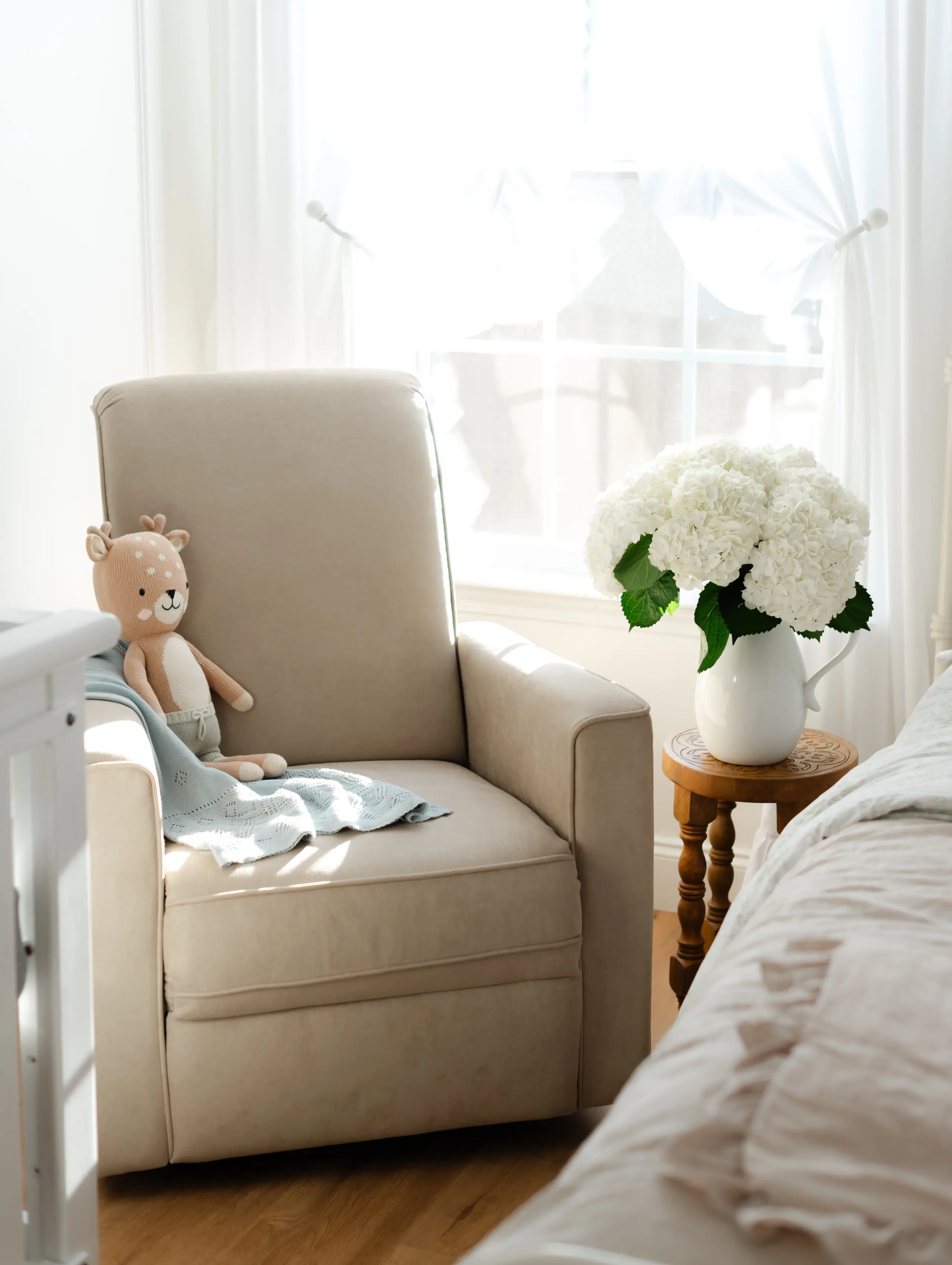 Evolur Raleigh glider recliner chair in fawn with stuffed toy and blanket beside bed and flowers in bright sunlit room