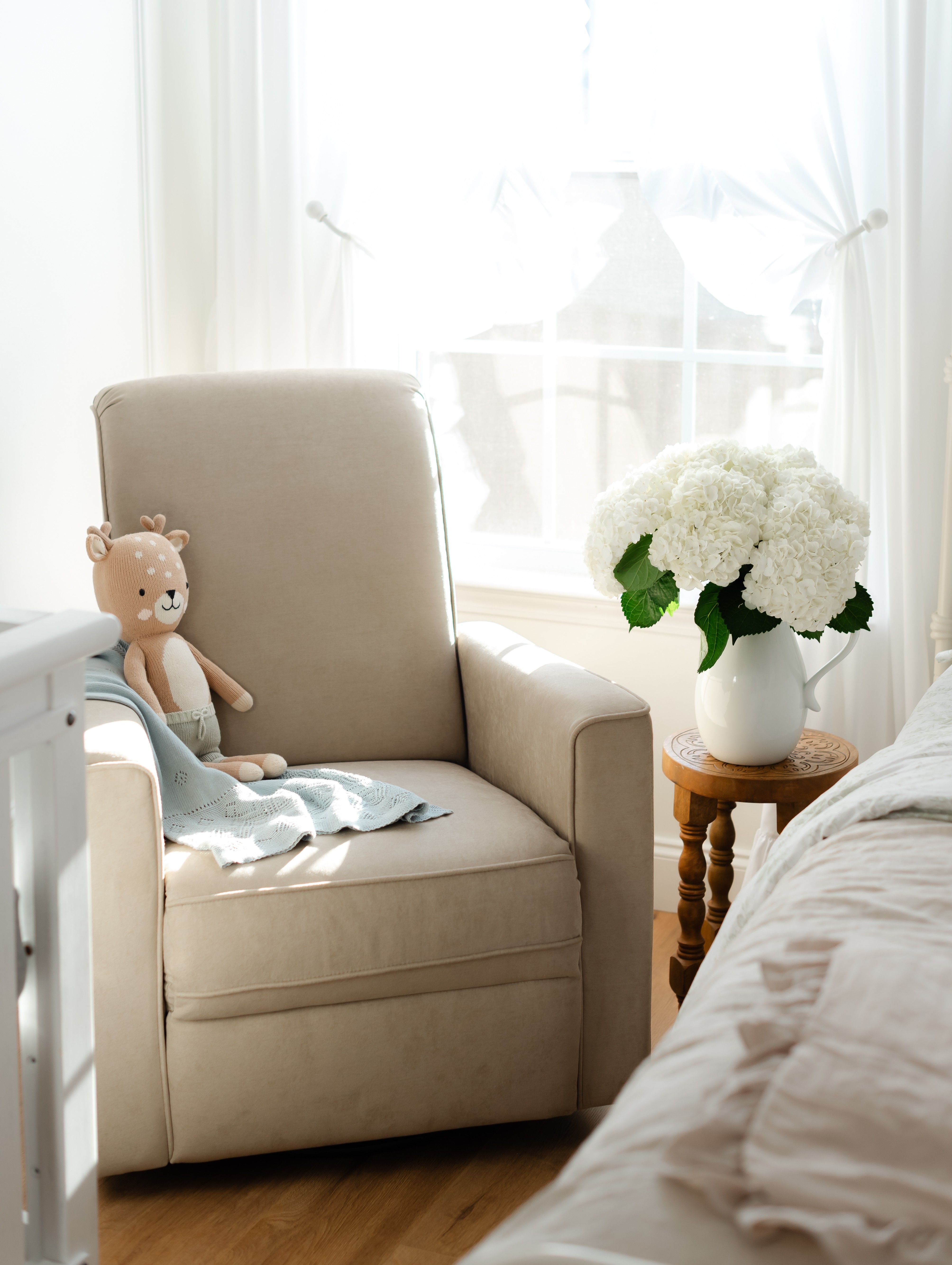 Evolur Raleigh glider recliner chair in fawn with stuffed toy and blanket beside bed and flowers in bright sunlit room
