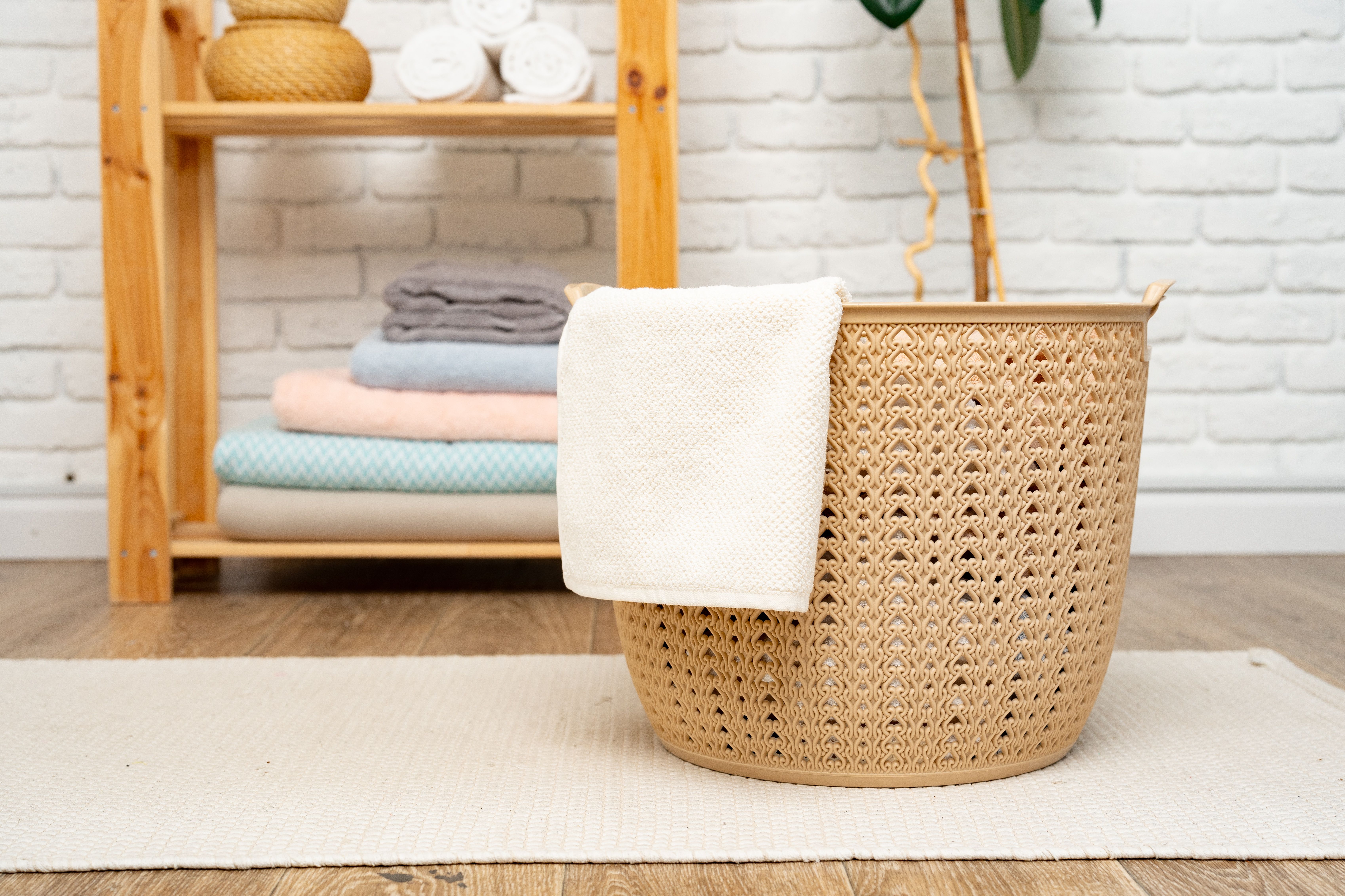 Beige woven-style plastic laundry basket with a white towel draped over the edge, placed on a cream rug in a cozy laundry room with neatly folded towels and wooden shelving in the background.