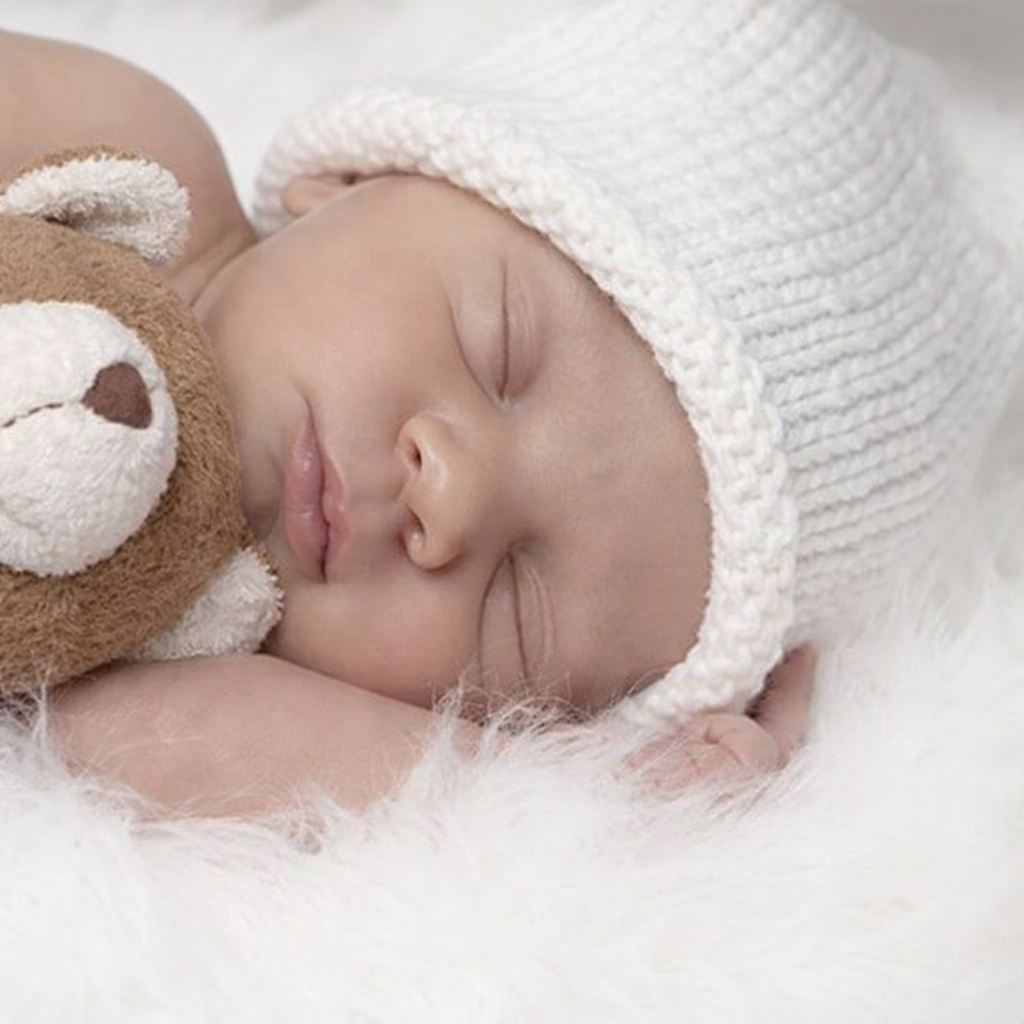 Baby sleeping wearing a soft knit hat, cuddling a plush teddy bear on a cozy white blanket