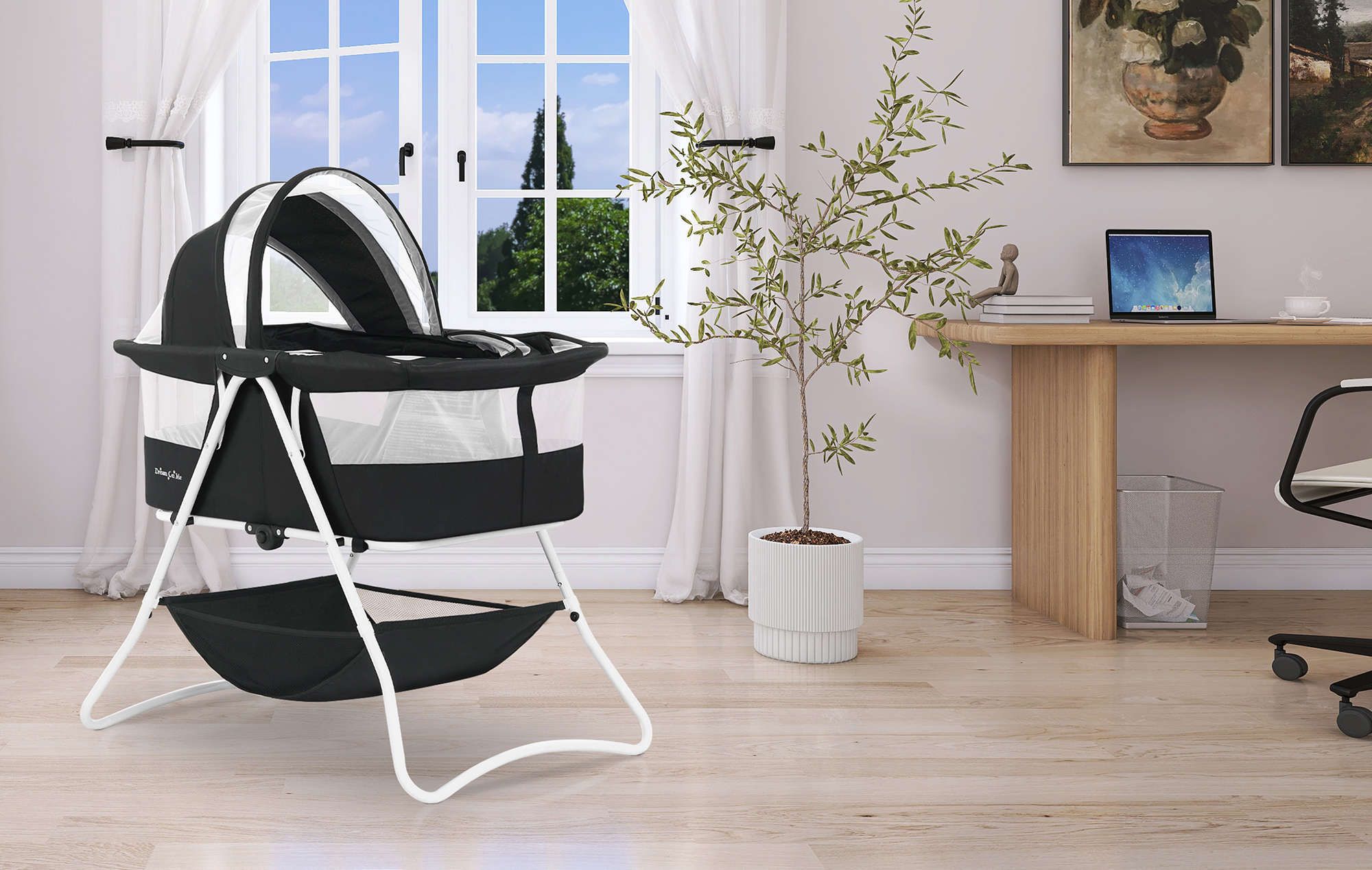 Dream On Me Karley bassinet with mesh sides and canopy, placed in a modern home office with a wooden desk, potted plant, and large window with white curtains