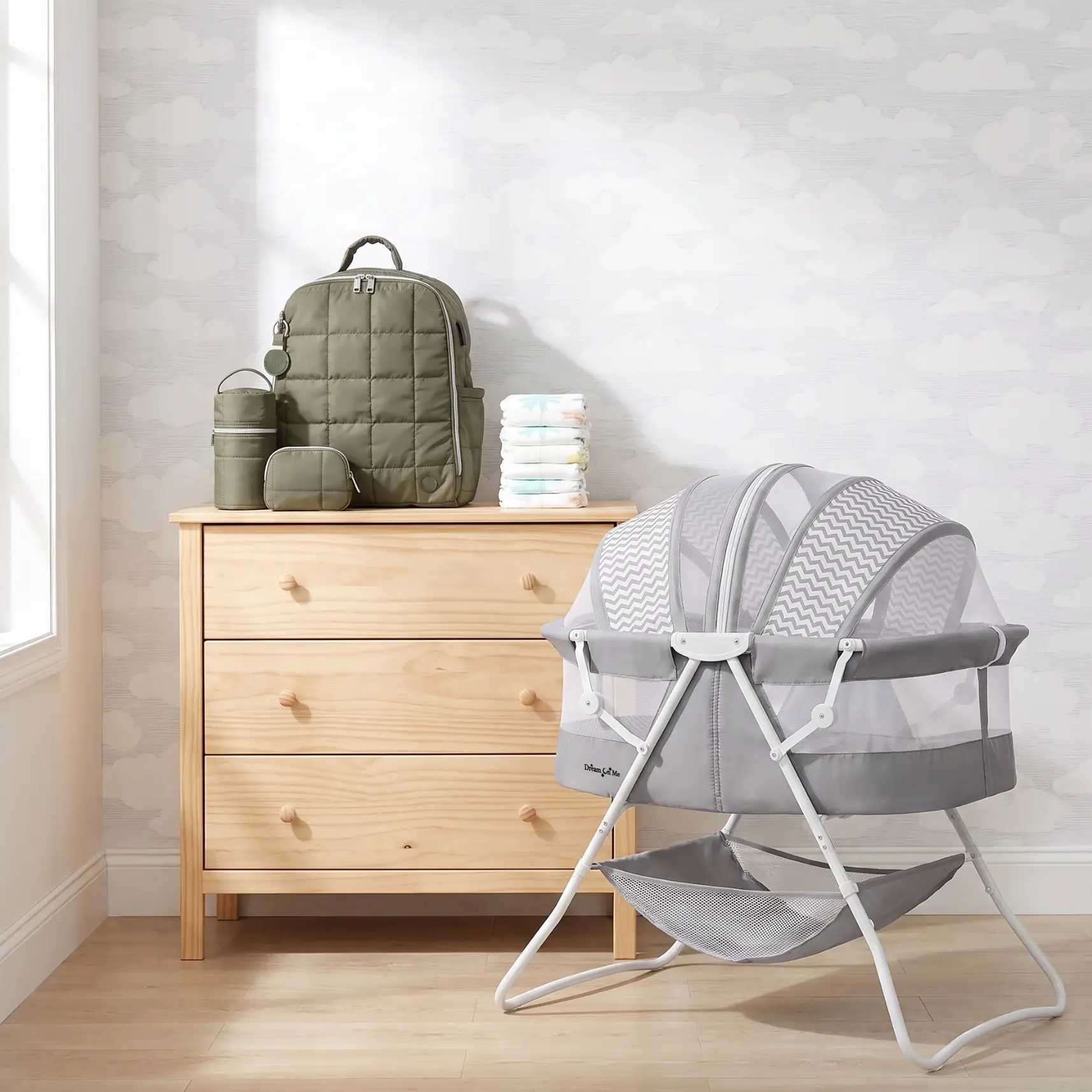 A wooden dresser topped with an olive green diaper bag set and diapers, next to a gray portable bassinet with a canopy.