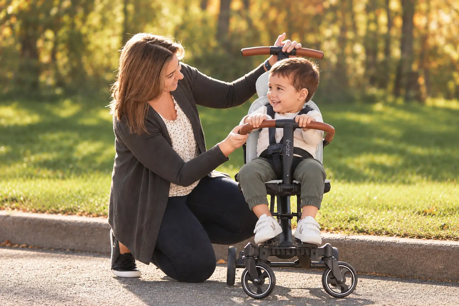 A woman smiles at a happy toddler in a gray push trike outdoors.