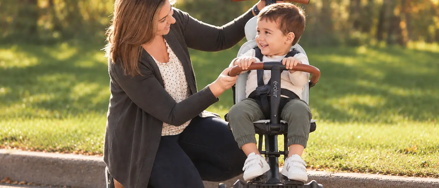 A woman smiles at a happy toddler in a gray push trike outdoors.