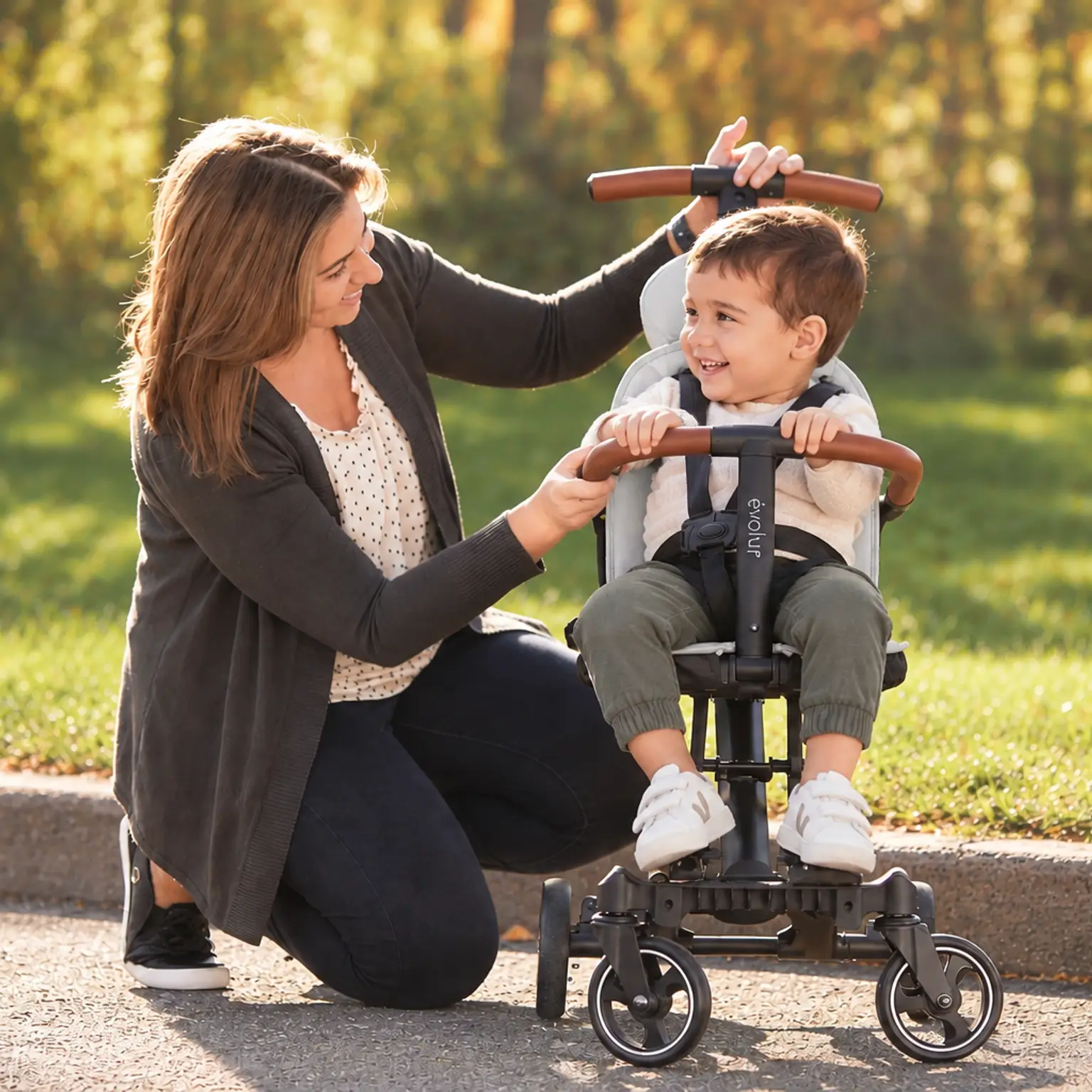 A woman smiles at a happy toddler in a gray push trike outdoors.