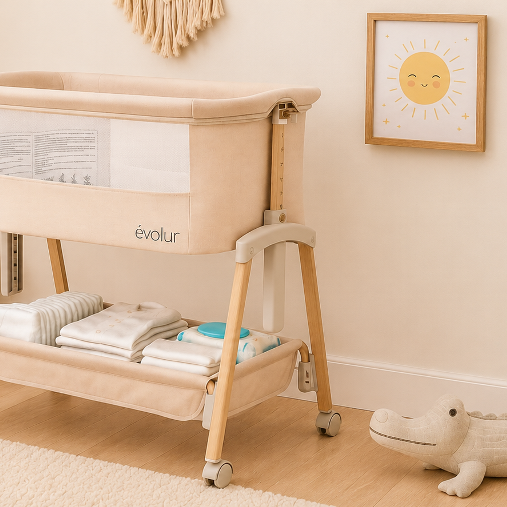 A beige Évolur baby bassinet with storage below, in a softly lit nursery with an alligator toy and sun picture.