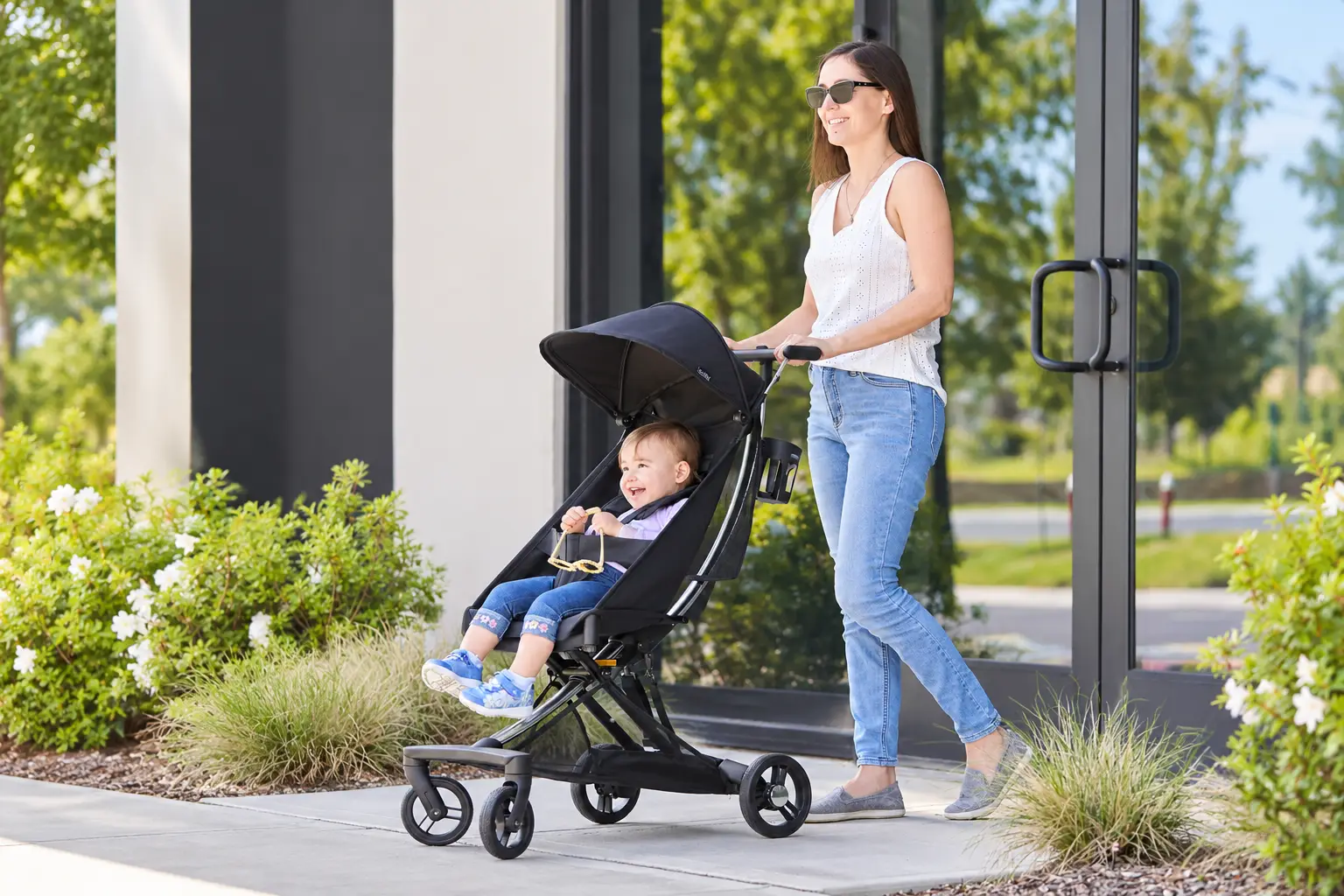 A smiling woman pushes a happy baby in a black stroller outdoors.