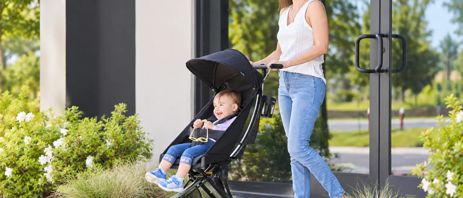 A smiling woman pushes a happy baby in a black stroller outdoors.