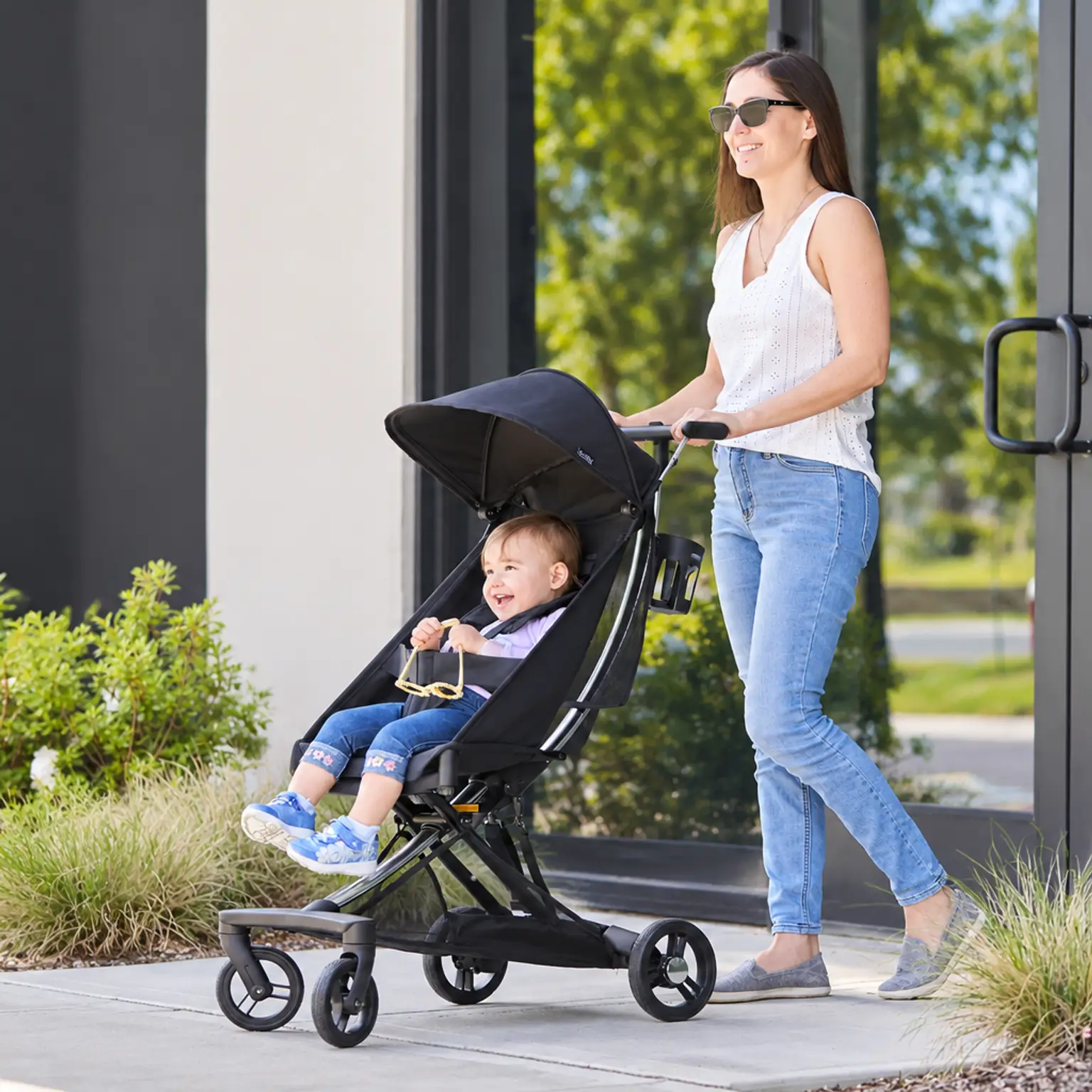 A smiling woman pushes a happy baby in a black stroller outdoors.