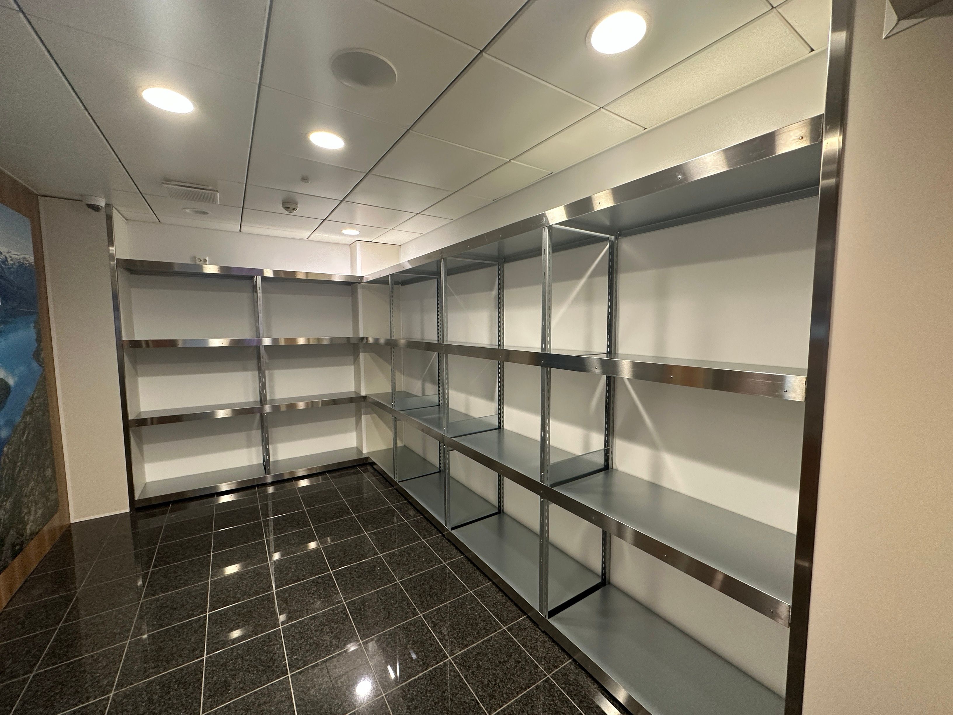An empty room with extensive L-shaped metal shelving along white walls and a dark tiled floor.