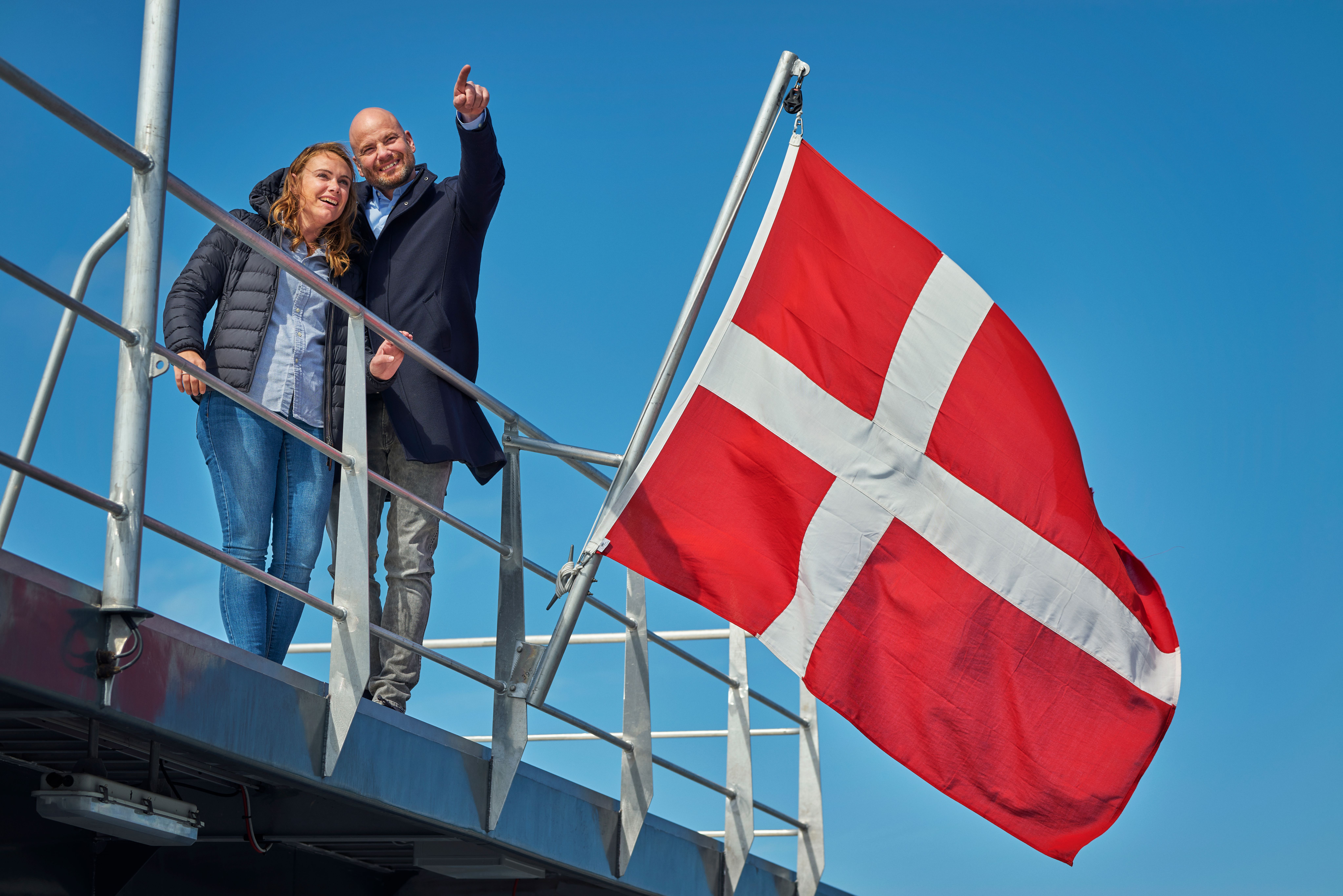 A man pointing and a woman standing on a deck next to a Danish flag.
