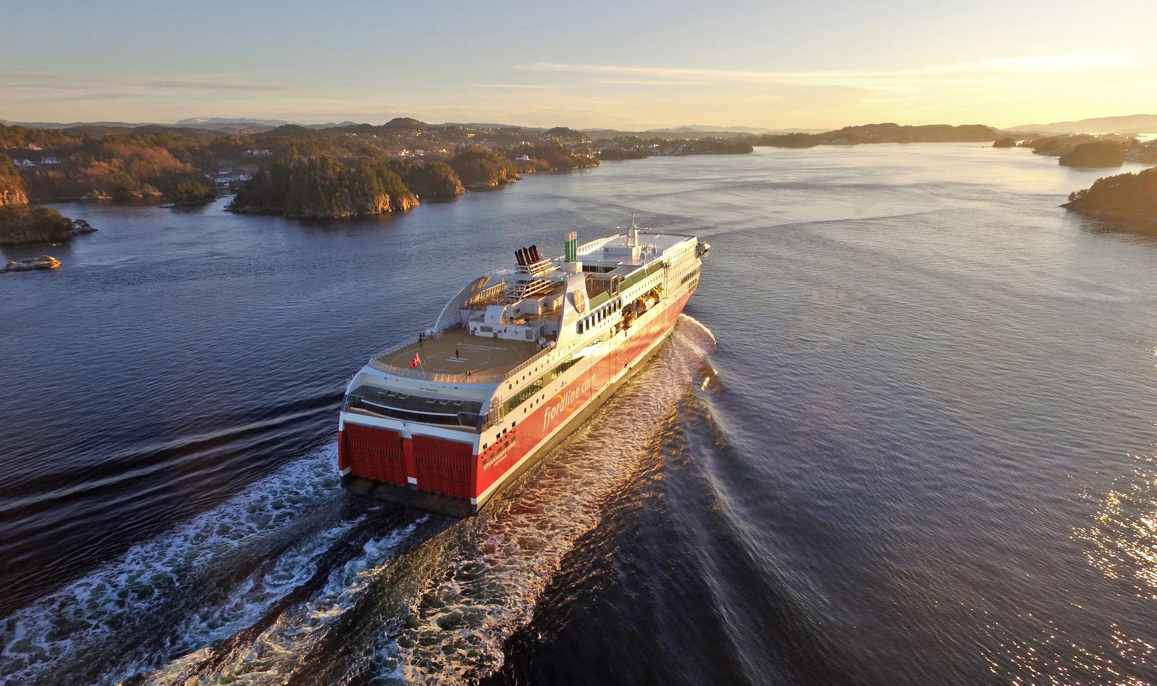 Eine Fjord Line-Fähre fährt bei Sonnenuntergang durch die norwegische Schärenlandschaft, im goldenen Licht.