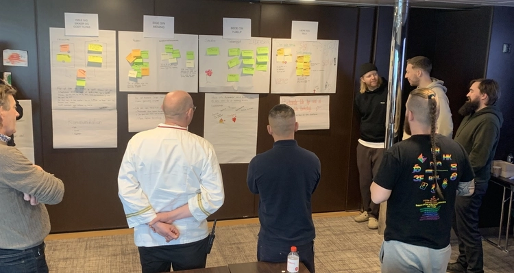 a group of men are standing around a table in front of a wall with sticky notes on it .