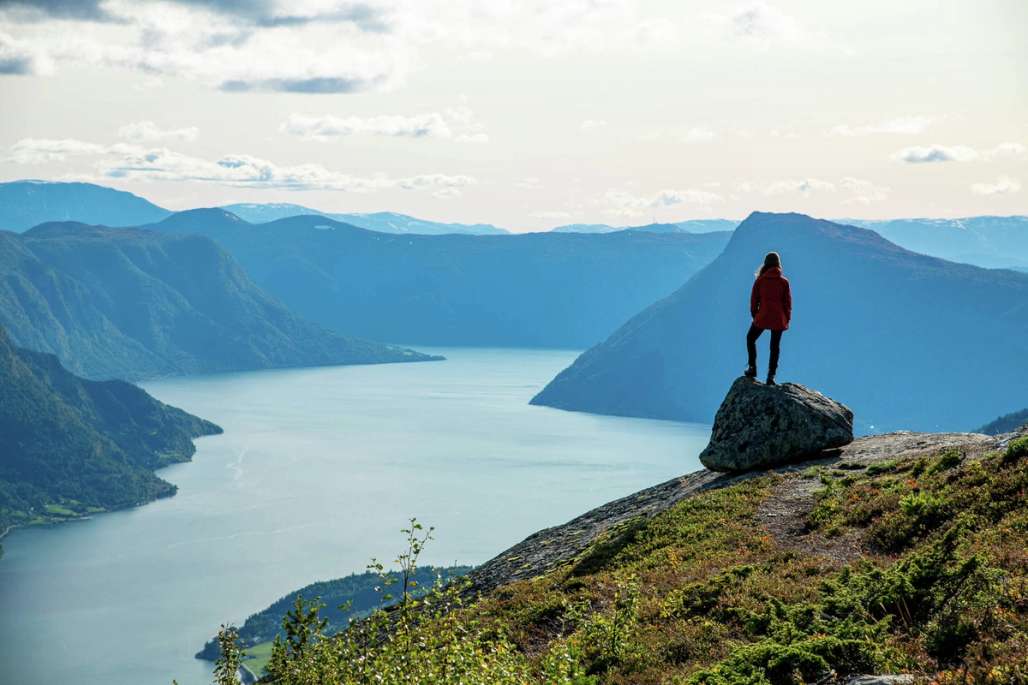 Eine Frau, die auf einem großen Felsen auf einem Berggipfel steht und über Berge und Fjorde blickt.