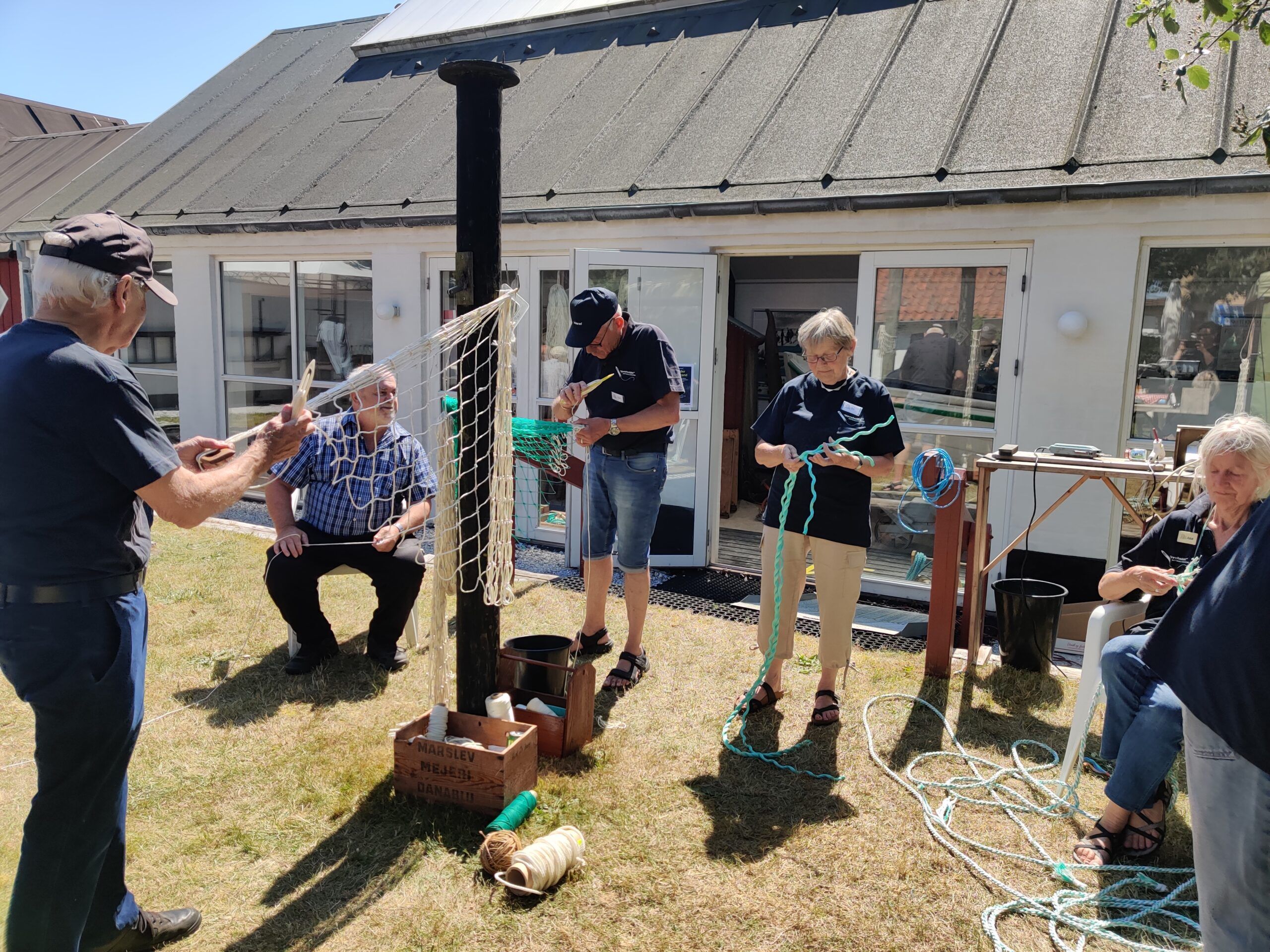 En gruppe eldre personer sitter og står rundt et nettverksted utendørs, hvor de jobber med tau og nett. Flere materialer og verktøy er plassert på gresset rundt dem.