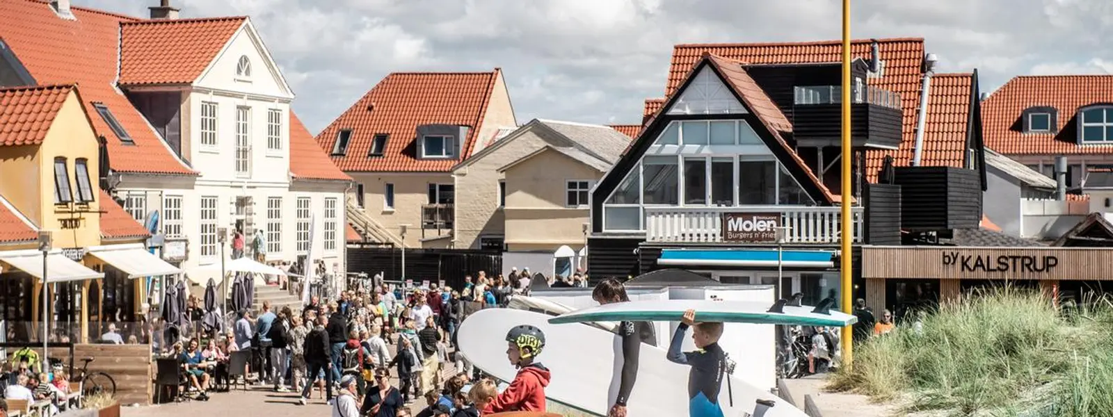 Et travelt torg med folk som går rundt, spiser på kafeer og handler. To personer med surfebrett går langs kanten av torget mot stranden.
