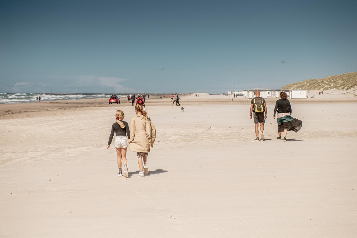 Flere personer går langs en bred sandstrand på en solrik dag, med sjøen og strandhytter i bakgrunnen.