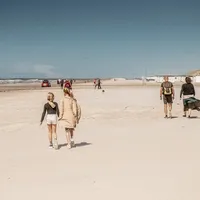 Flere personer går langs en bred sandstrand på en solrik dag, med sjøen og strandhytter i bakgrunnen.