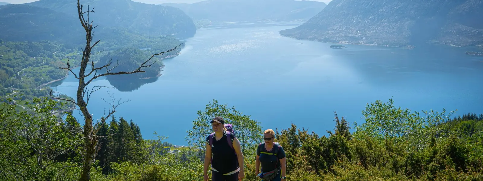 To damer på tur i grønne omgivelser med fjord og fjeld i baggrunden.
