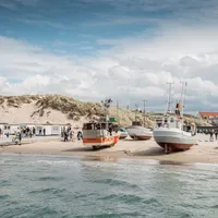 En kystscene med fiskebåter trukket opp på stranden foran sanddyner, med folk som spaserer og nyter dagen. Det er små hytter langs stranden og en blå himmel med noen skyer.