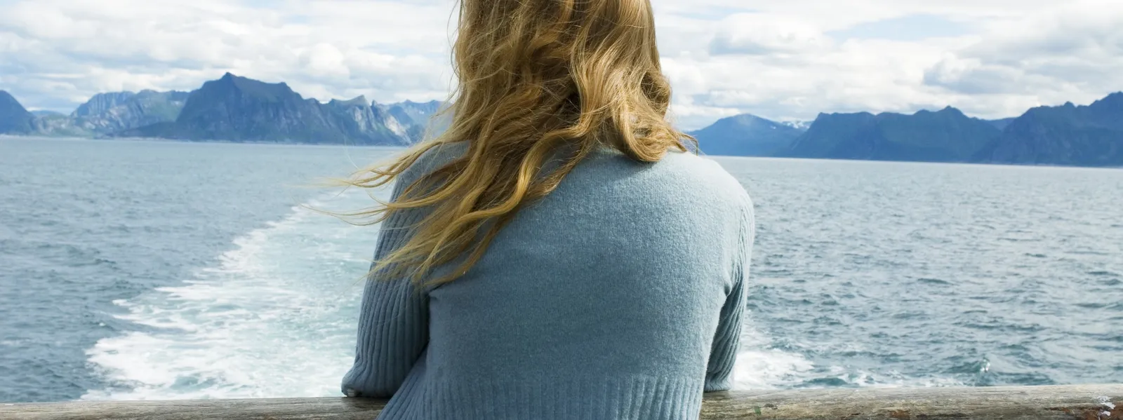 A woman stands in a boat and looks out over the fjord and the mountains.