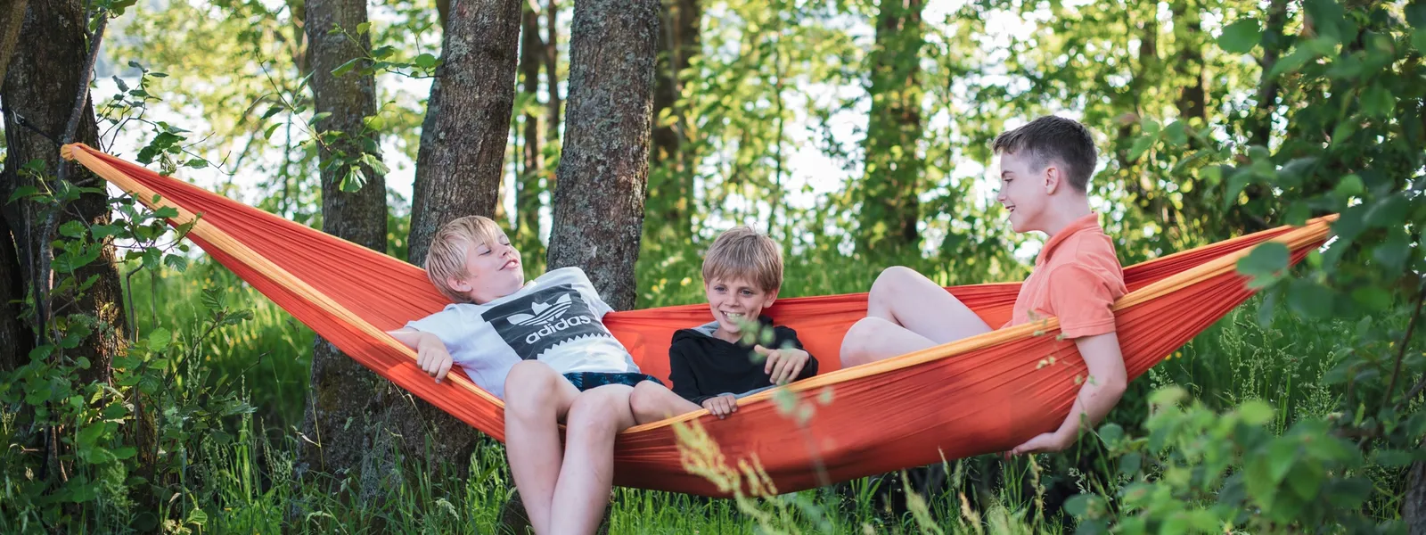 Three boys relaxing in an orange hammock hung between trees in a green wooded area.