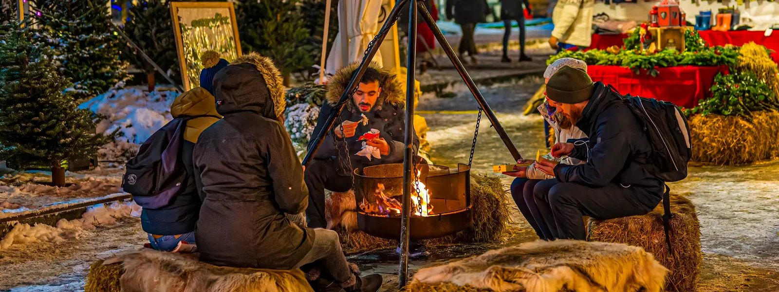 Mennesker sidder omkring et bål på et hyggeligt julemarked omgivet af juledekorationer og sne.