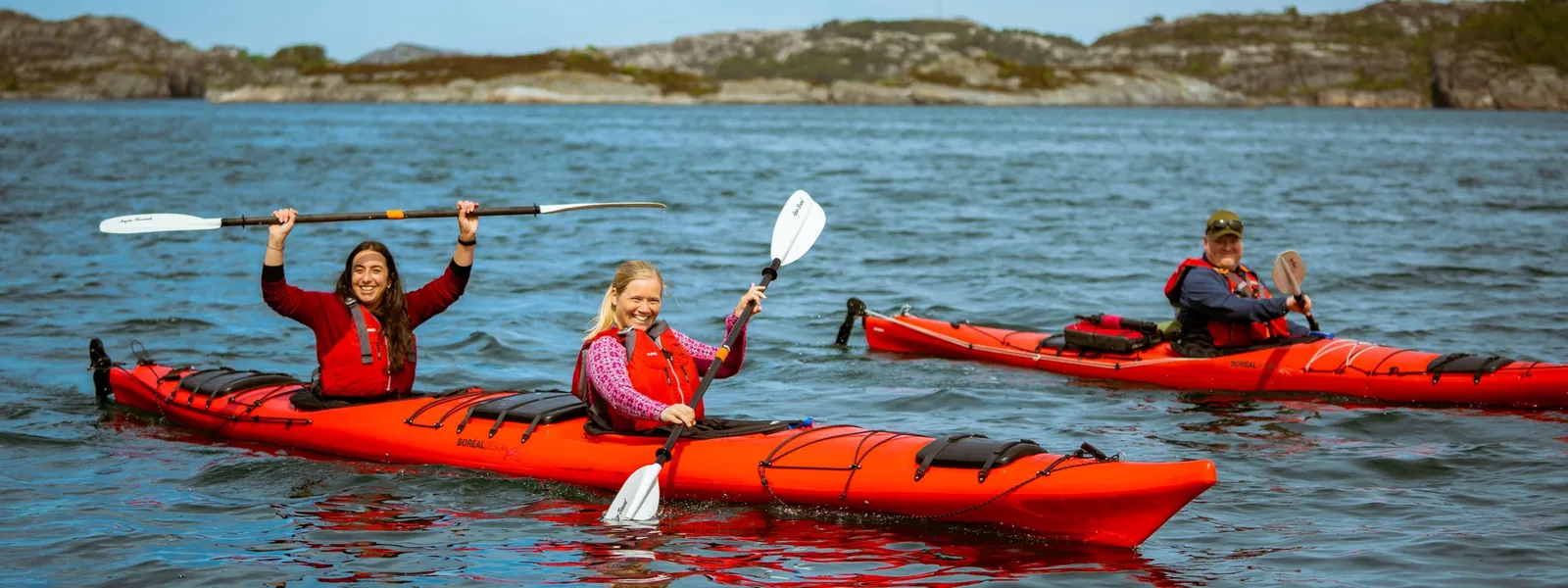 Three people kayaking on calm waters in red kayaks, enjoying an outdoor adventure on a sunny day.