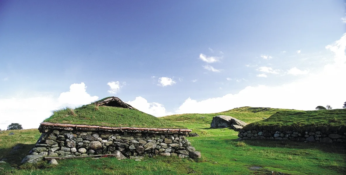 A traditional grass-covered stone house in a green landscape under a blue sky.