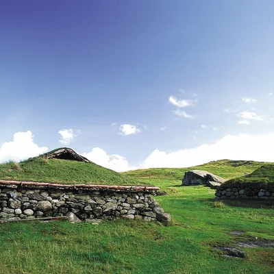 A traditional grass-covered stone house in a green landscape under a blue sky.
