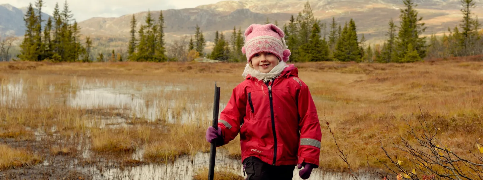 Et barn iført en rød jakke og en lyserød hue står med en stav i hånden i et sumpet fjeldlandskab om efteråret.