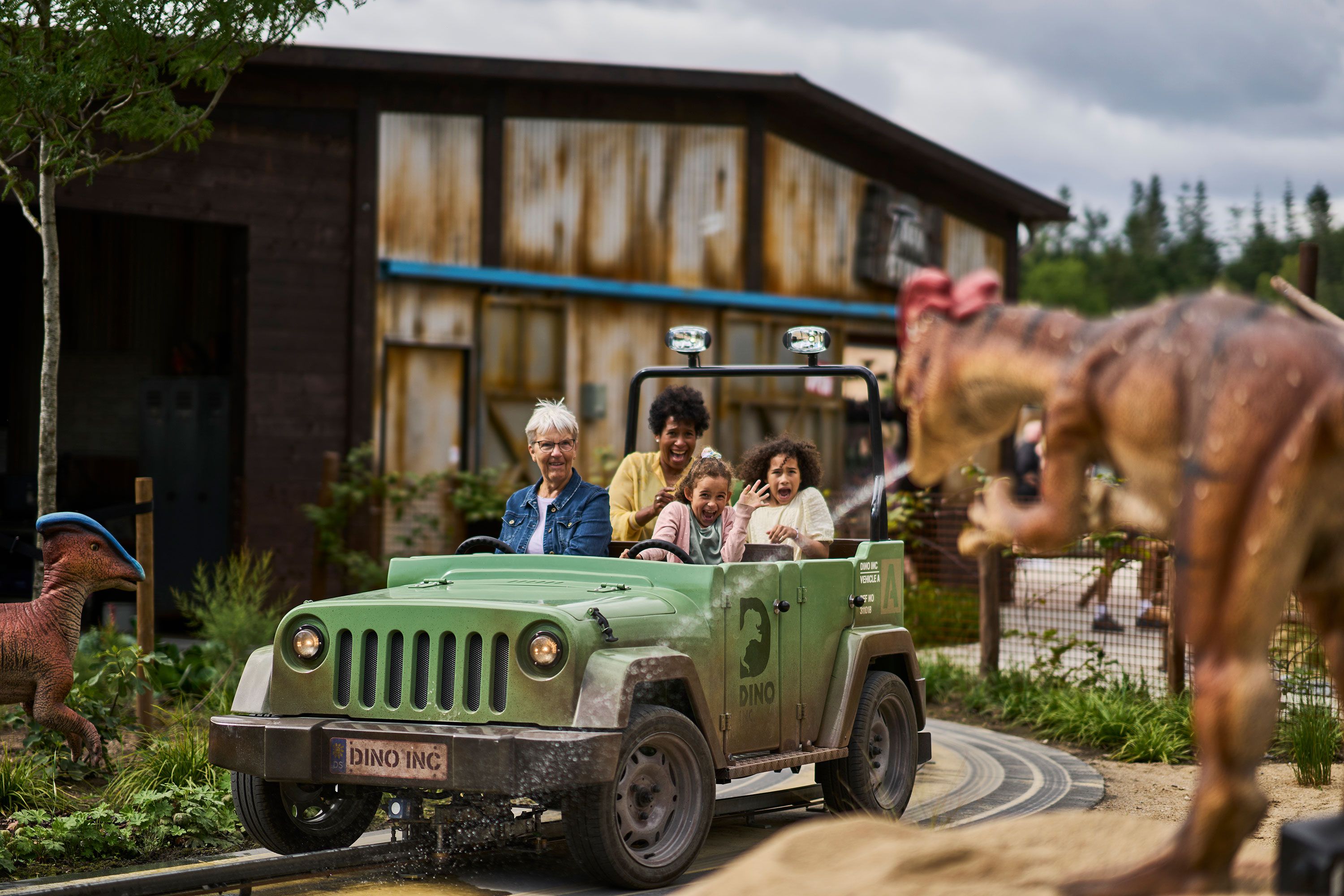 En familie på fire sitter i en grønn jeep på en fornøyelsesparkattraksjon med dinosaurtema, omgitt av dinosaurfigurer, med en rustikk bygning i bakgrunnen.