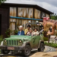 En familie på fire sitter i en grønn jeep på en fornøyelsesparkattraksjon med dinosaurtema, omgitt av dinosaurfigurer, med en rustikk bygning i bakgrunnen.
