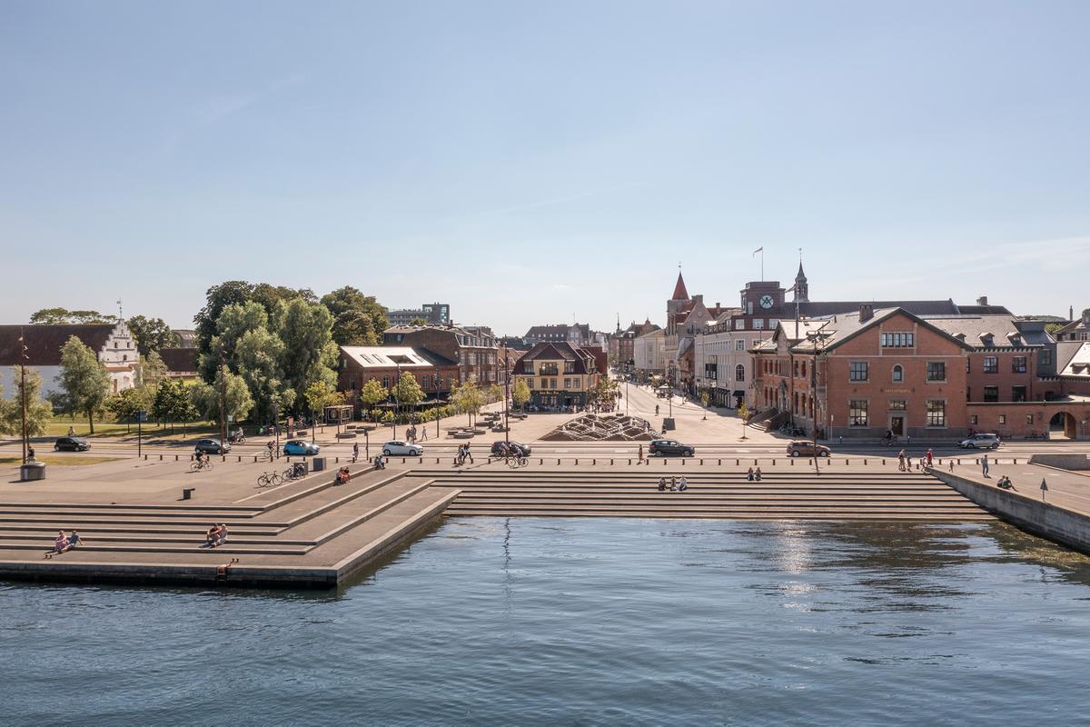 Utsikt over en bypromenade ved vannet med trapper ned til vannkanten, trær og historiske bygninger under en klar blå himmel.