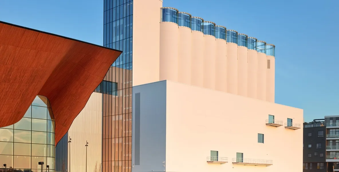 Moderne Gebäude an der Hafenfront bei Sonnenuntergang, mit einer markanten holzverkleideten Struktur links und einem hohen, weißen Gebäude rechts.