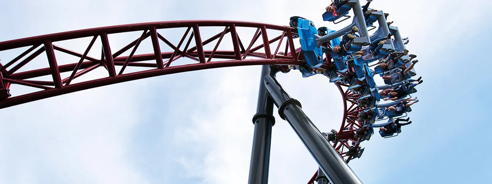 En berg-og-dal-bane med røde vogner fylt med mennesker som smiler og roper mens de kjører nedover en høy bue, under en blå himmel med noen skyer.