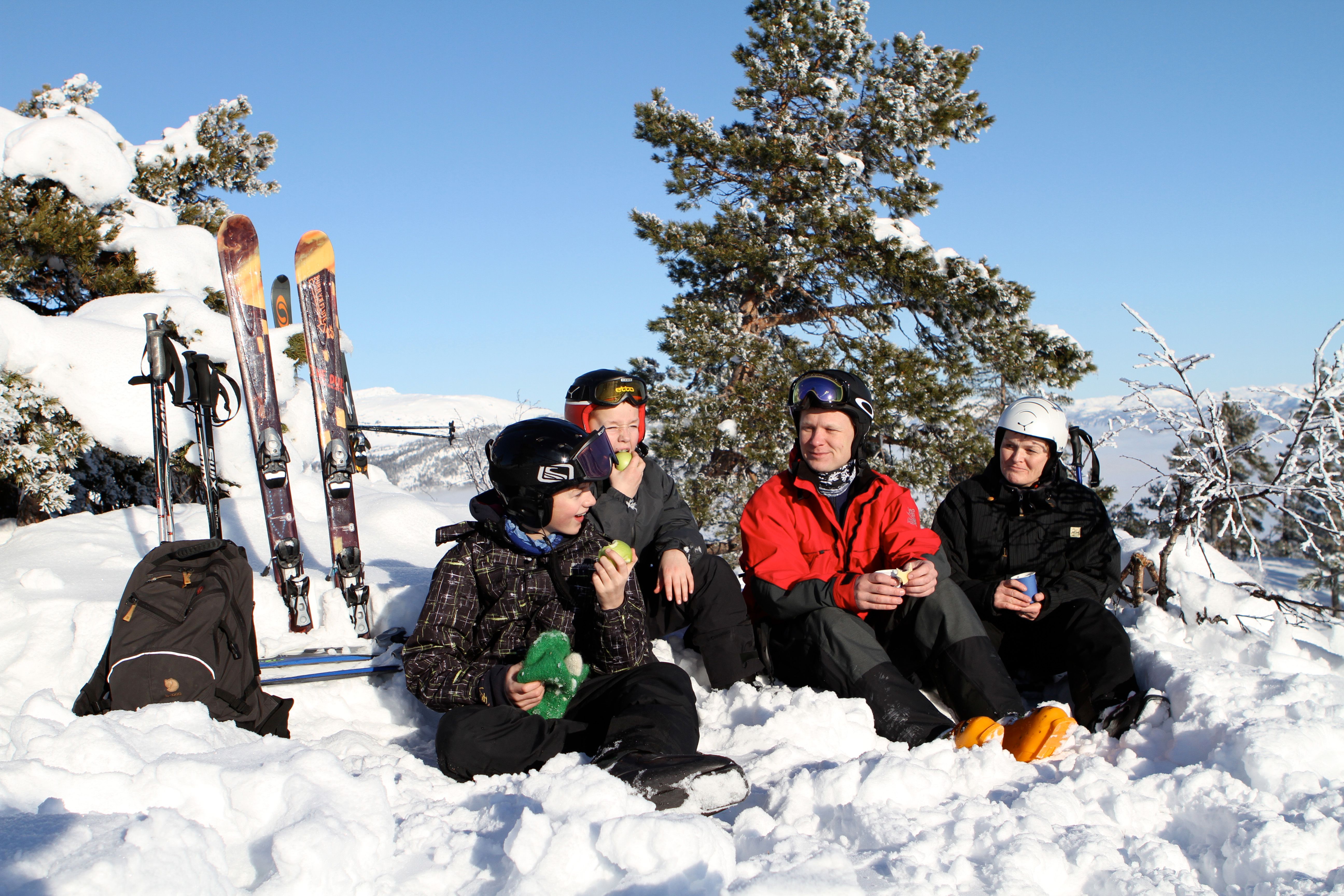 En skifamilie holder pause i sneen og nyder snacks under den klare blå himmel.