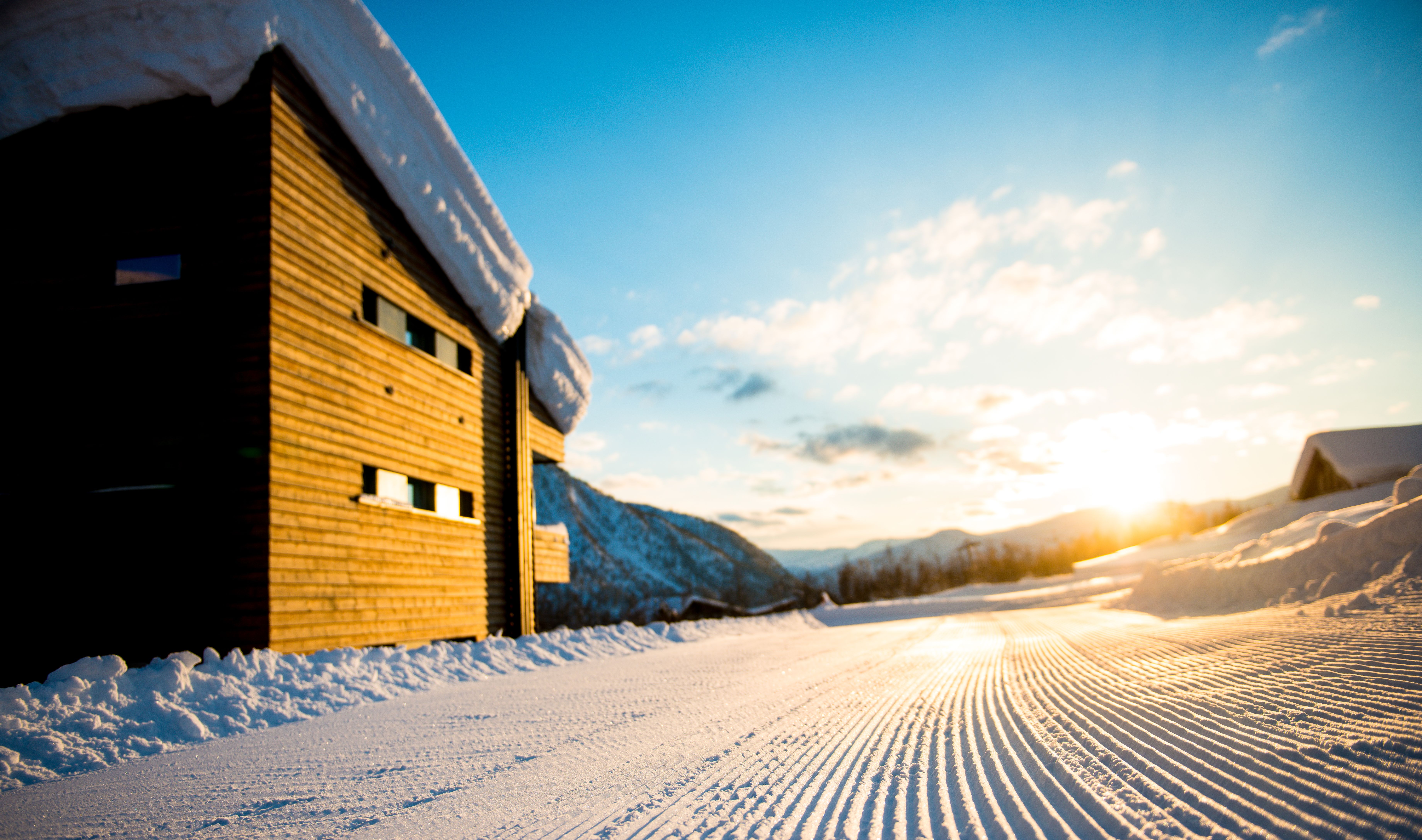 A wooden cabin covered in snow sits alongside freshly groomed snow with the sun rising in the background.