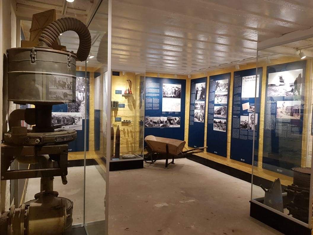 Utstilling i et museum med historiske gjenstander fra andre verdenskrig, inkludert en gammel maskin, verktøy og informasjonstavler med bilder og tekst på veggene. En trillebår og andre utstilte gjenstander er også synlige.