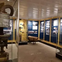 Utstilling i et museum med historiske gjenstander fra andre verdenskrig, inkludert en gammel maskin, verktøy og informasjonstavler med bilder og tekst på veggene. En trillebår og andre utstilte gjenstander er også synlige.