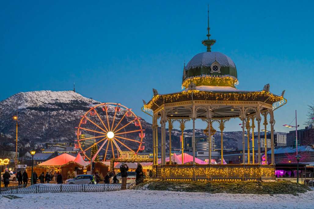 Weihnachtslichter und ein Riesenrad erhellen einen Winterabend in Bergen, mit schneebedeckten Bergen im Hintergrund.